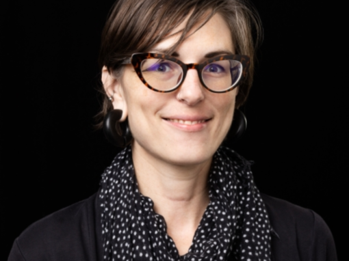 Portrait of a smiling woman wearing tortoiseshell glasses and a patterned black scarf against a dark background.